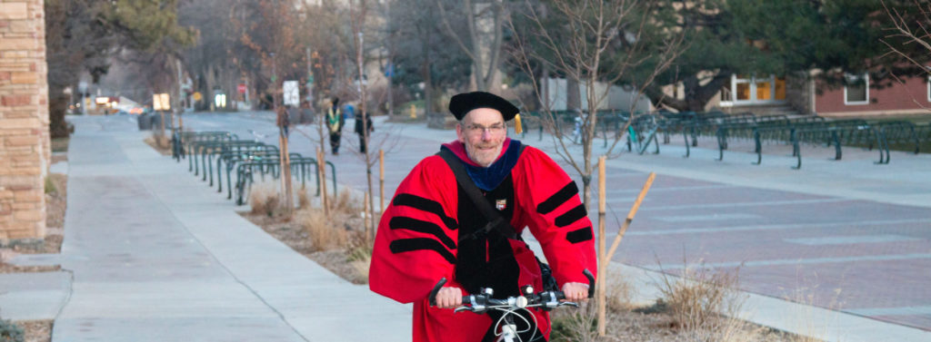 Dean John Hayes riding a bike with his commencment regalia on