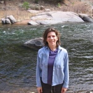 Dr. Stephanie Kampf stands on a riverbank and smiles toward the camera. The river is behind her. 