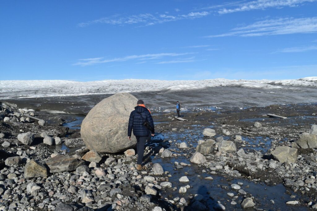 A man walks through a rock field next to a giant boulder. 