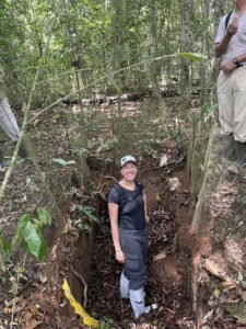 A woman stands in a soil pit in a tropical rainforest to conduct research on root systems.