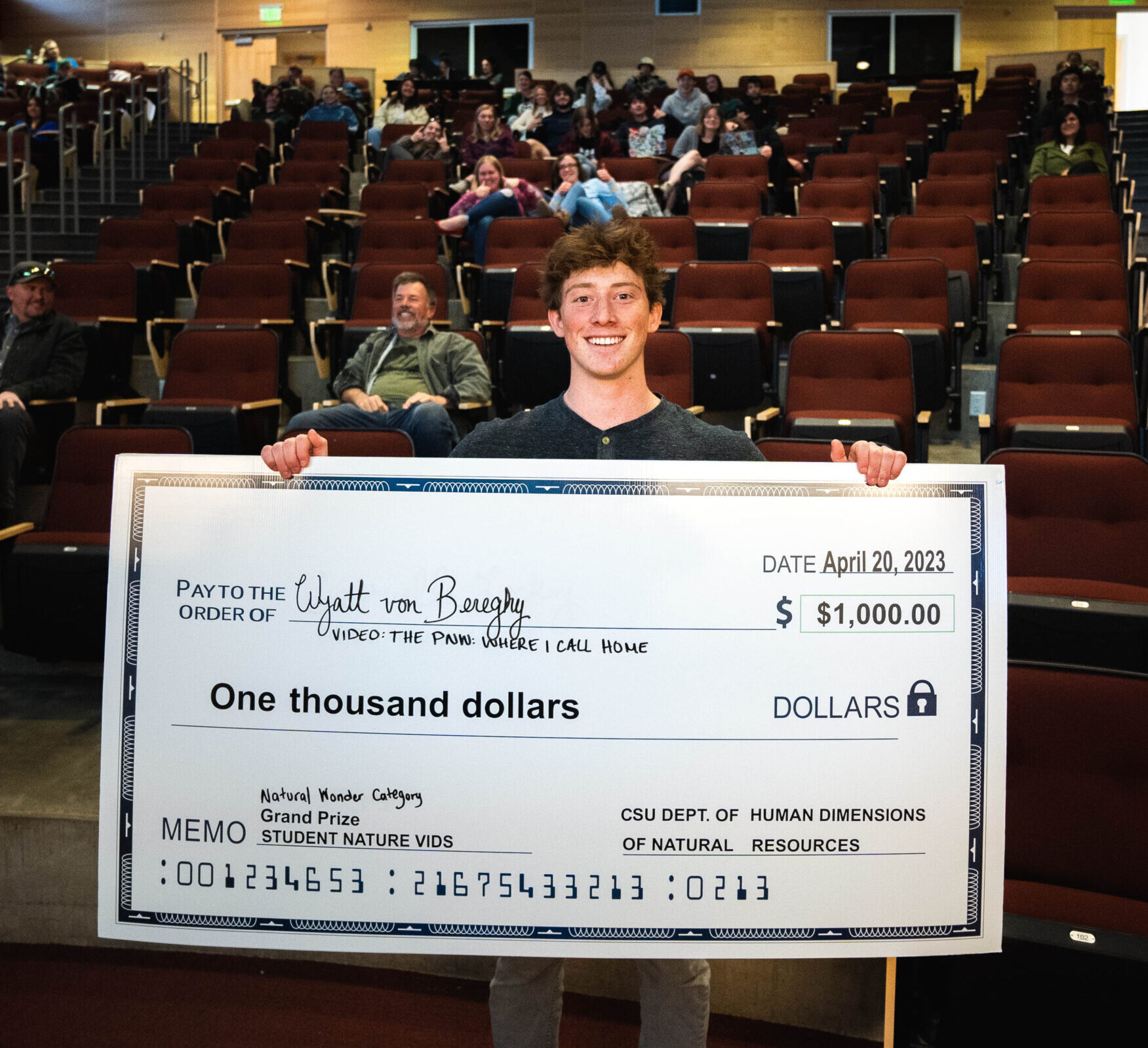 A man stands holding a large $1,000 check in an auditorium.