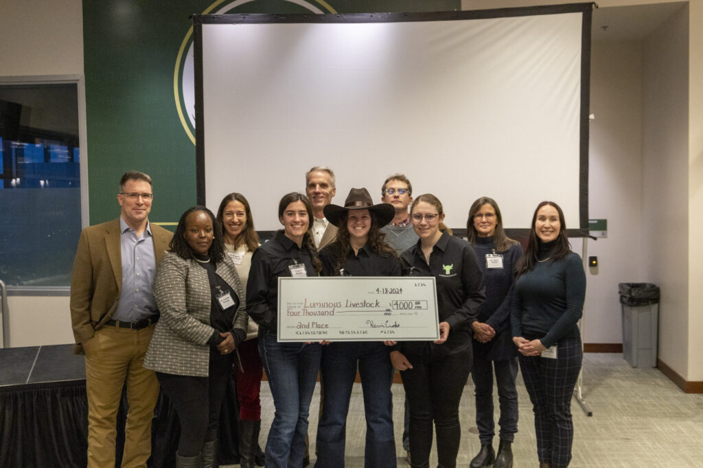 A group of College students and faculty stand in front of a screen, holding a large check for the amount of $4,000 in prize winnings.