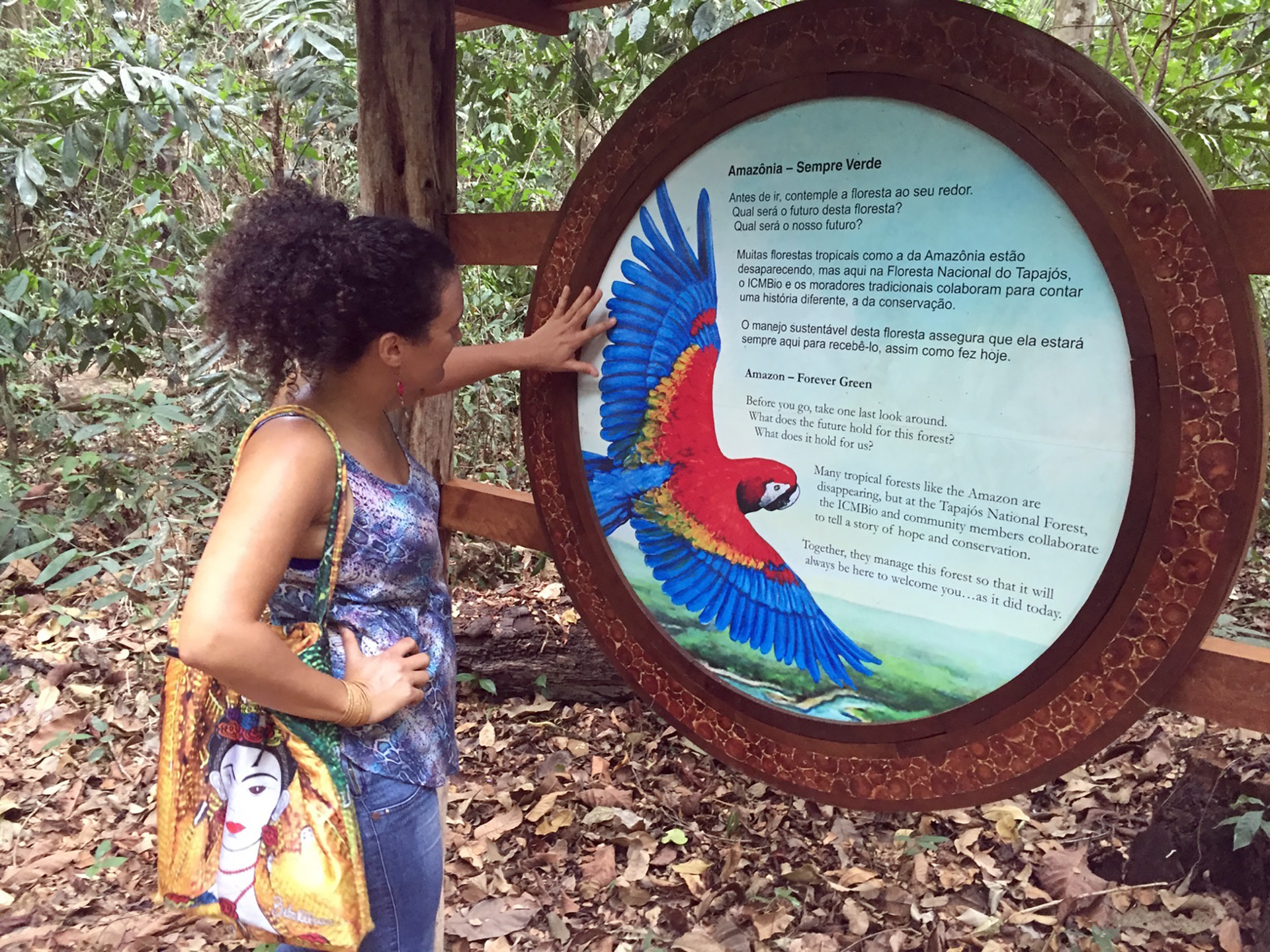 A woman touches the feathers of a colorful tropical bird painted on a sign at a park in an Amazon forest.