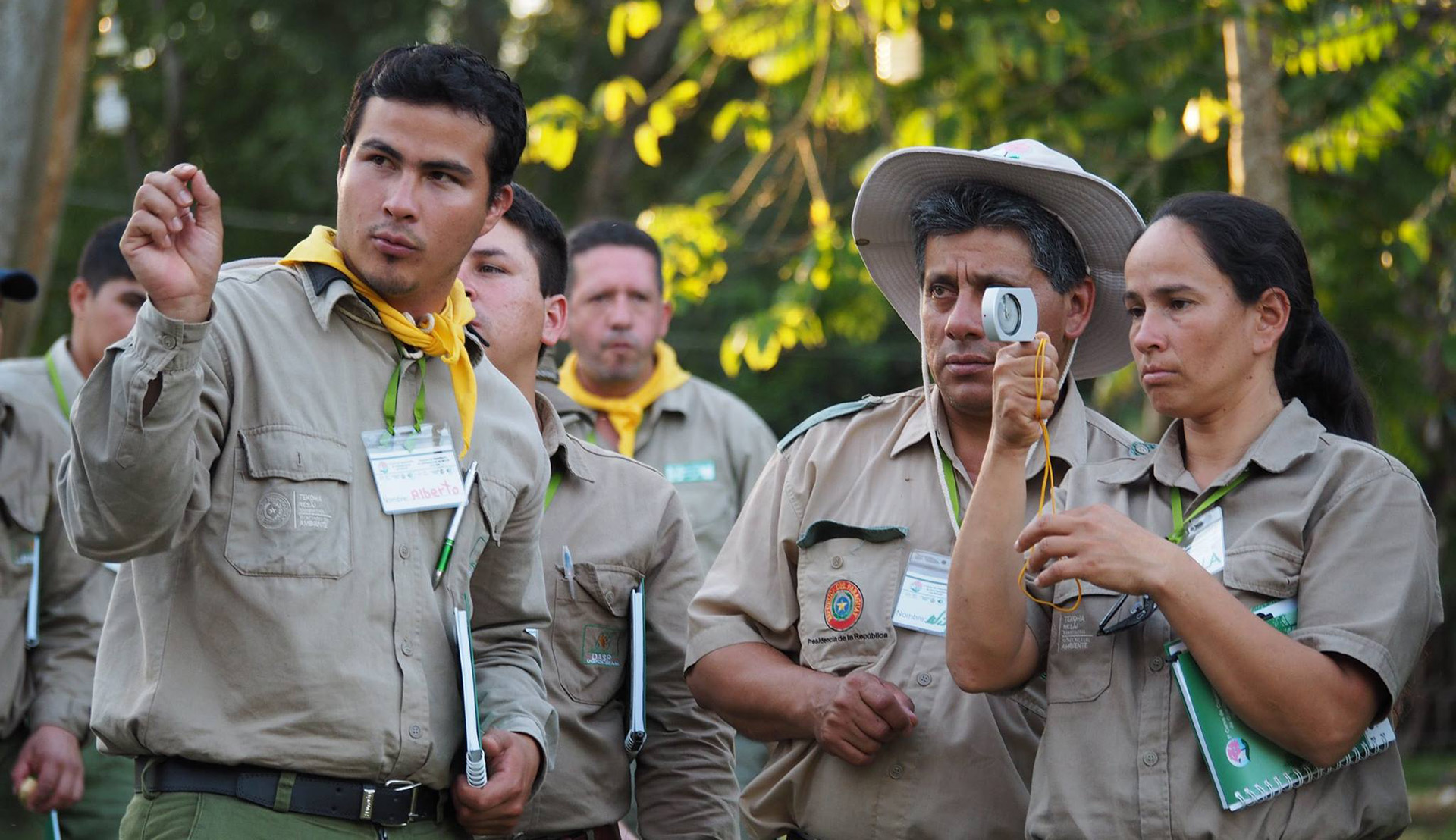 A group of people in ranger uniforms stands in a forest; all hold notebooks and one holds a measurement instrument.