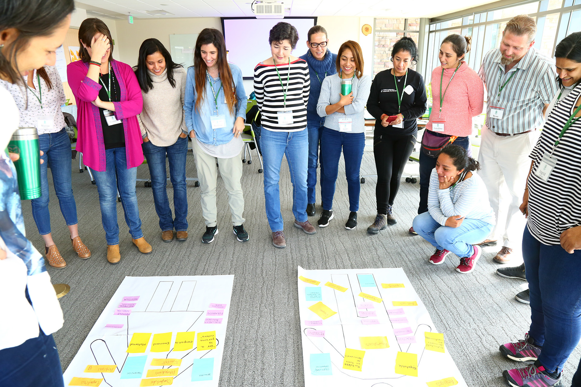 A group of smiling women and one man stand around and look down at two posters on the floor of a meeting room. The posters have outlines representing a man and woman on them and are covered in colored post-it notes with writing.