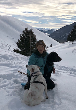 Hanna McCaslin, in a teal jacket, smiles while sitting in snow-covered mountains with two dogs, one white and one black, beside her. Snowy peaks and overcast skies are visible in the background.