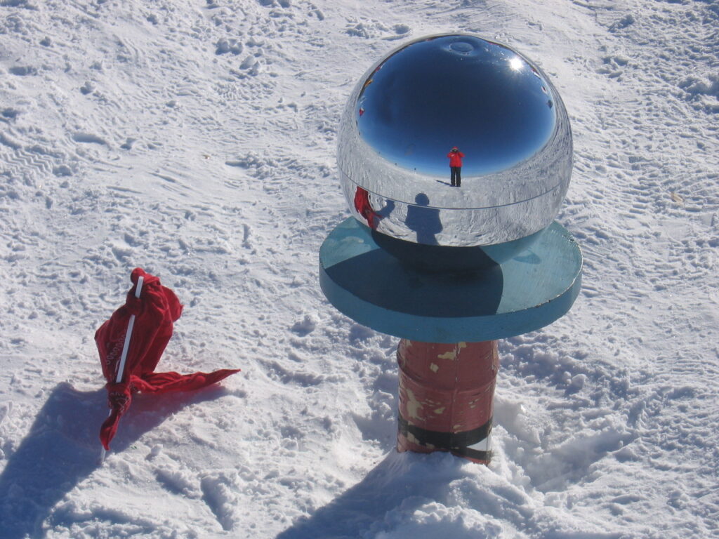 A round seismic instrument sits in the snow, with a reflection of a man (the photographer) wearing a red snow suit on the surface.