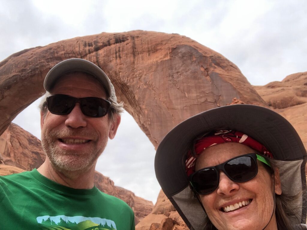 A man and a woman wearing hats and sunglasses take a selfie in front of a large rock formation in the shape of an arch.